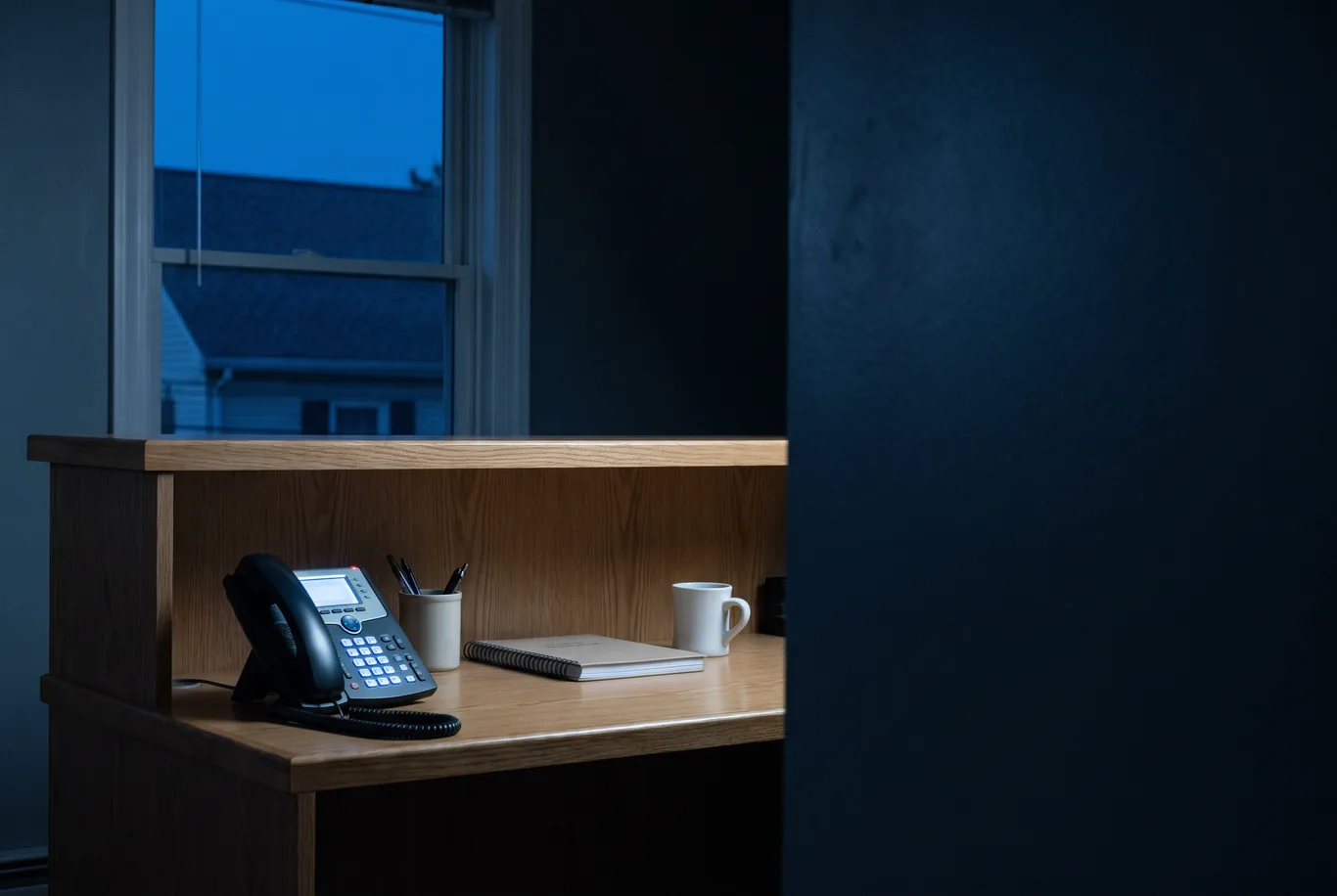 An empty reception desk at dusk with a desk phone, its keypad glowing, and a dim window behind it