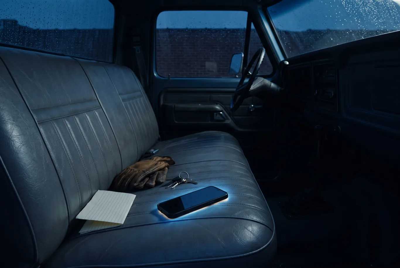 A phone lies on a truck bench seat at night with a glow around it, beside work gloves, keys and a blank note pad