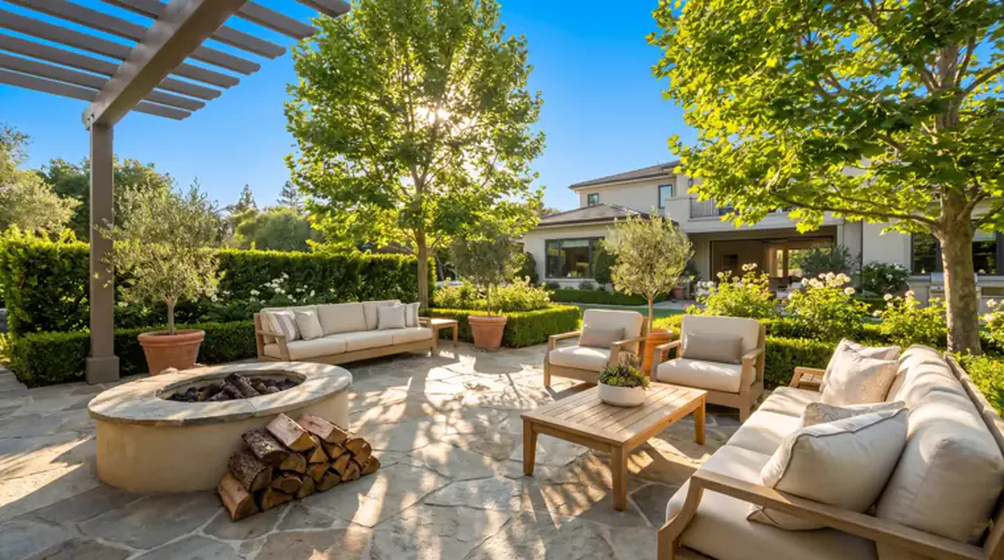 A flagstone patio in afternoon sun with a round stone fire pit, teak seating, clipped hedges and a pergola