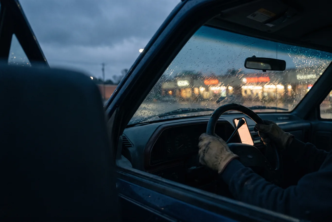 Inside a pickup cab at night, a phone lighting up in a work-worn hand, rain on the windshield behind it