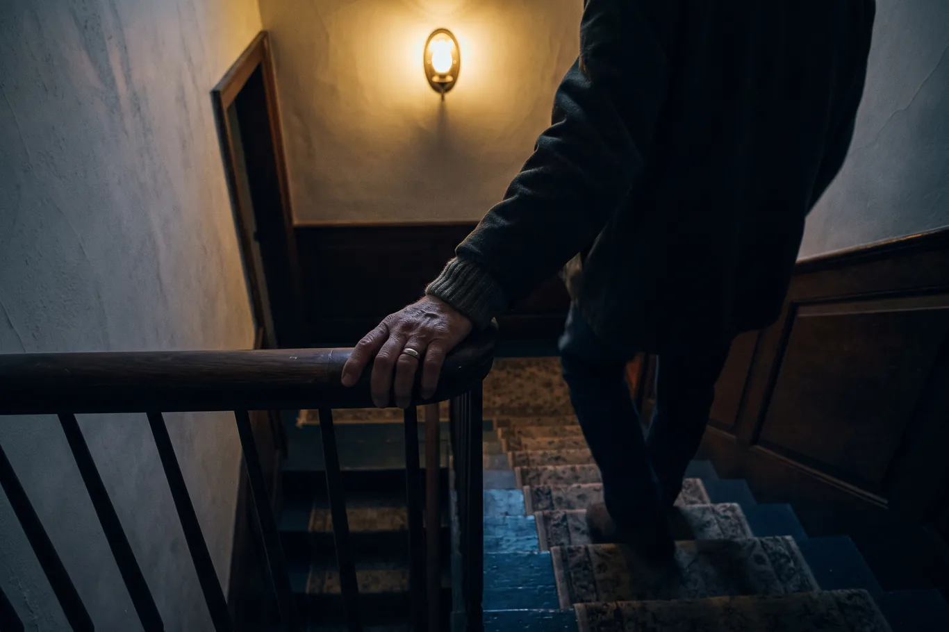 A hand gripping a stair rail in a dim colonial stairwell at night, weight shifted onto one leg