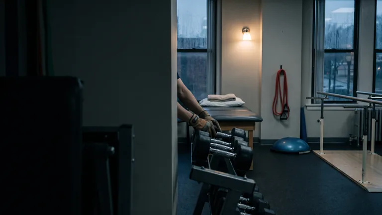 A physical therapy gym at dusk, hands re-racking a dumbbell on the rack beside a treatment table and a resistance band