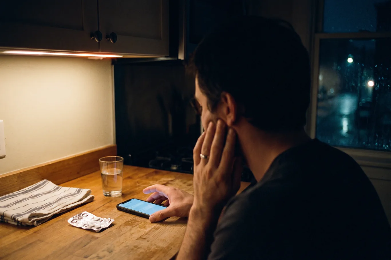 A patient's hand on a phone at a kitchen counter at night, one hand pressed to a jaw