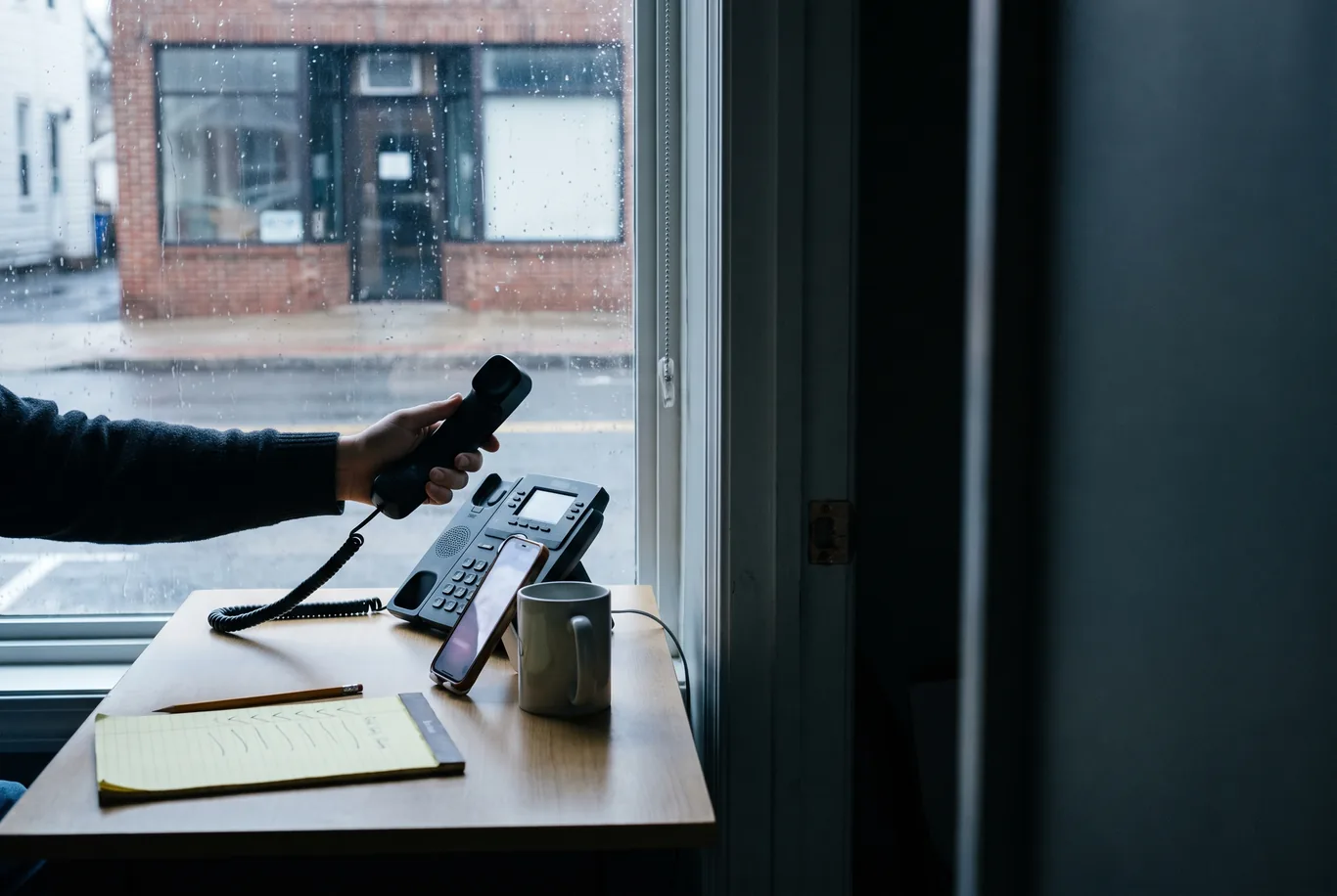 A desk by a rainy window on an overcast morning, a phone propped against a mug, a legal pad of pencil check marks, and a hand lifting a desk telephone handset
