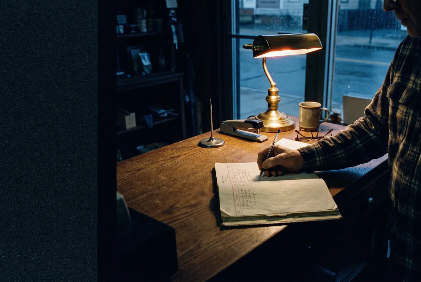 A hand writing in a paper ledger book on a shop counter at closing time, one desk lamp on and the shop dark behind