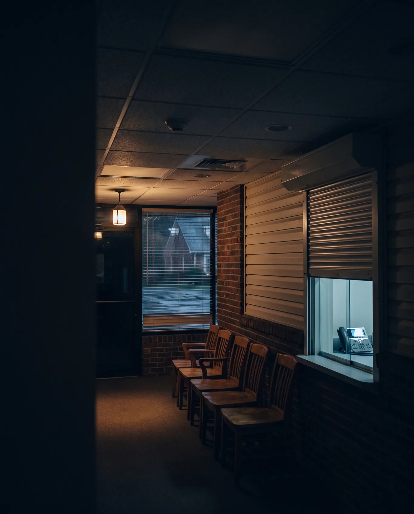 A medical office waiting room after hours, chairs in a row, the reception window's shutter half down, a phone's glow behind it