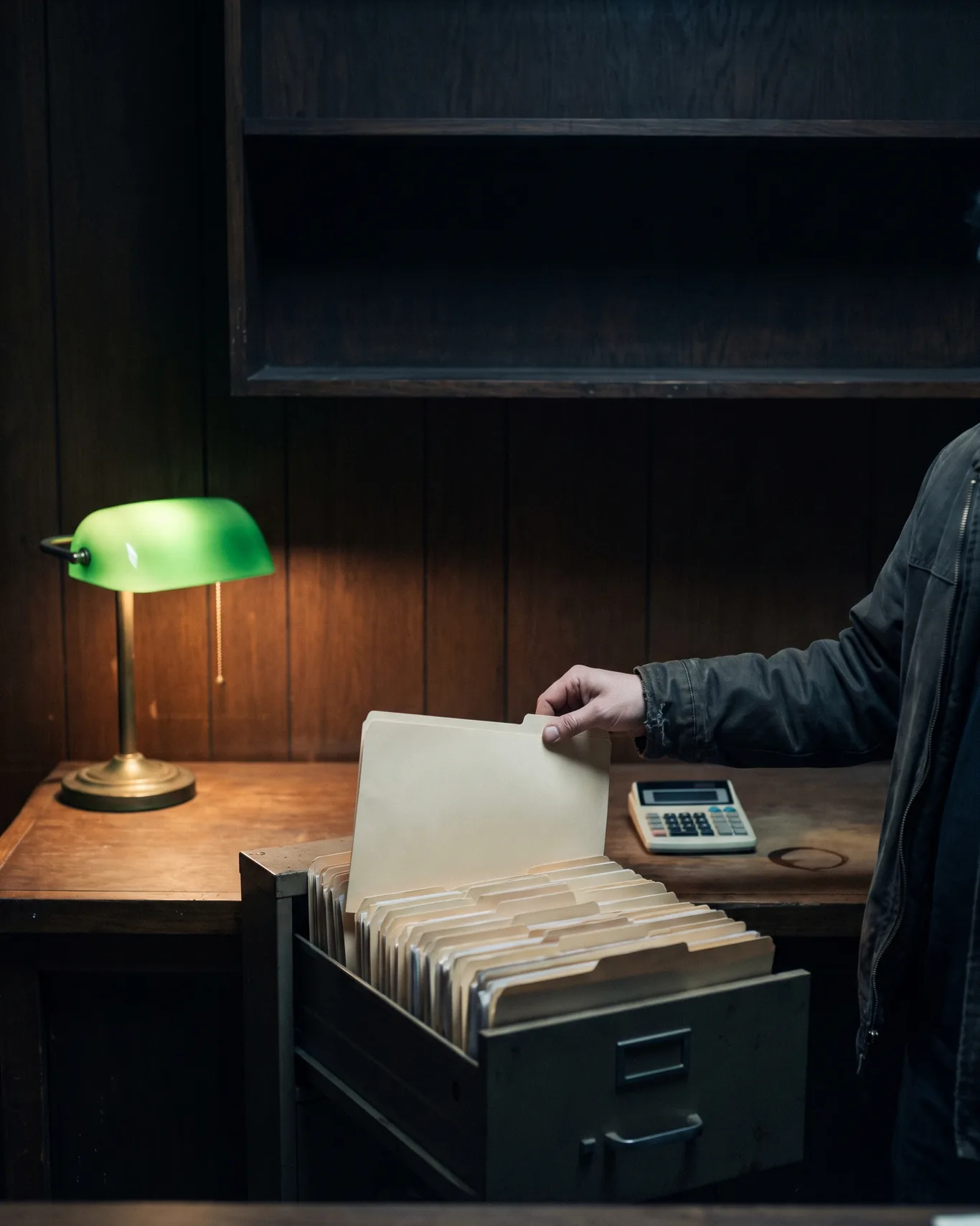 A hand pulling one folder from a drawer of old estimate folders on a shop counter under a lamp