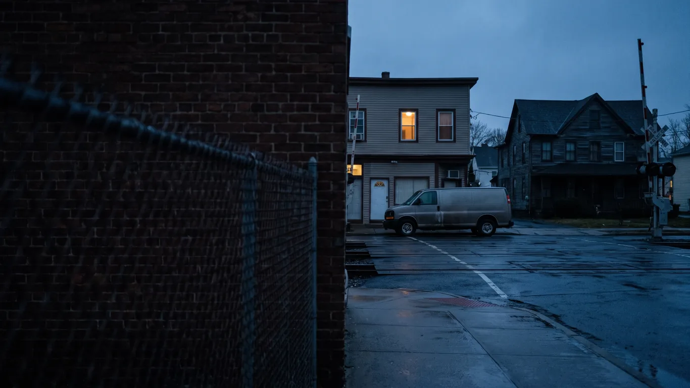 A low borough avenue at blue hour with a freight rail crossing and a work van at the kerb, wet asphalt, no readable signs