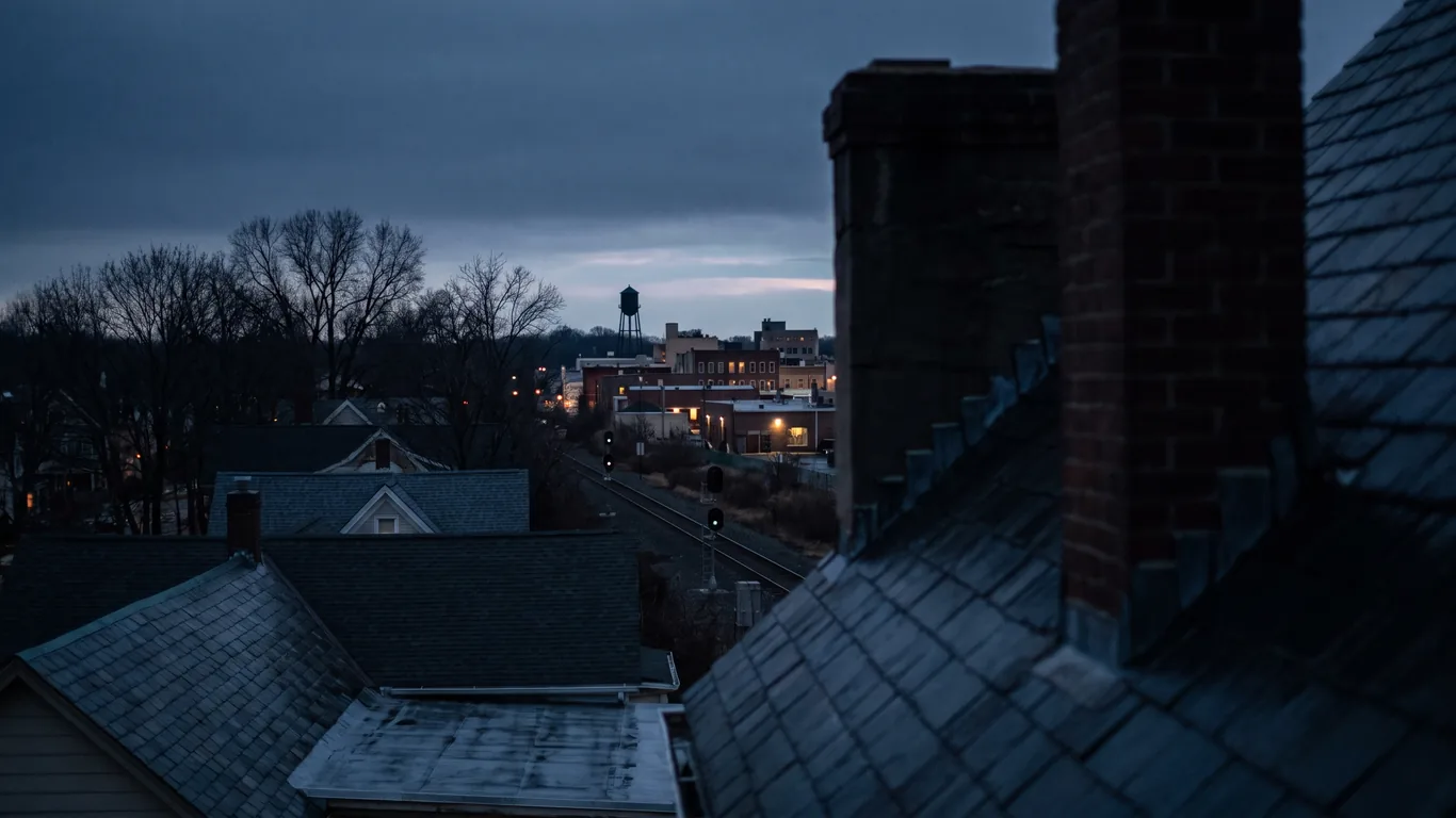 A wide view over rooftops and a rail line toward distant downtown lights at blue hour, wet slate roofs and bare trees